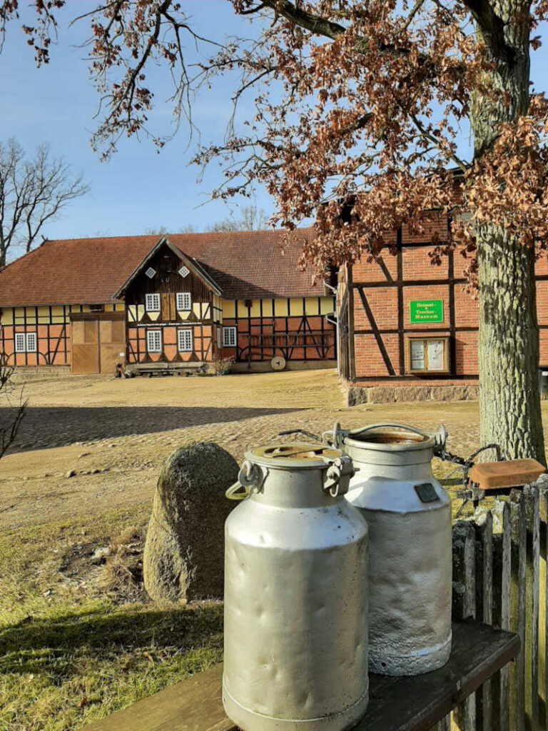 Im Vordergrund zwei Milchkannen und im Hintergrund der Hof Scharnhop mit dem Heimat-& Treckermuseum Niendorf I